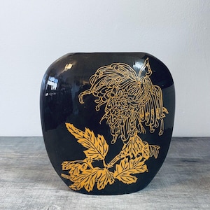 May include: Black ceramic vase with a gold floral design. The vase has a rounded shape and features a stylized chrysanthemum and leaves.