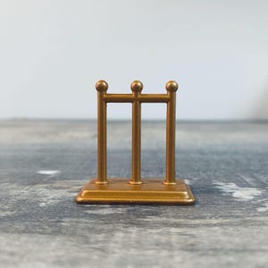 May include: A small, gold-colored decorative stand with three vertical bars and a rectangular base. The top of each bar has a small, round ball. The stand is made of a shiny, metallic material.