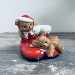 May include: A ceramic Christmas decoration featuring two brown teddy bears. One bear wears a Santa hat and peeks out of a red boot with white trim. The other bear lies on the boot. The boot has a black sole.
