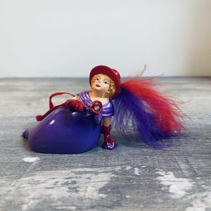 May include: A figurine of a woman in a purple dress and red hat, gloves, and shoes. The figurine has a red and purple feather plume and a jewel on the chest. The figurine is lying down.