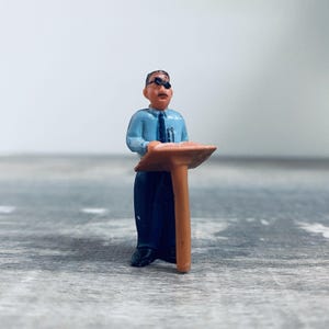 May include: A miniature figurine of a man in a light blue shirt, dark blue pants, and black shoes. He wears sunglasses and stands behind a brown podium. A pen is in his shirt pocket. The figurine is on a gray surface.