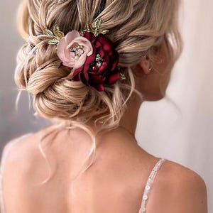 May include: A hair accessory featuring two fabric flowers in blush pink and deep burgundy, accented with gold-toned metal leaves and small clear crystals. The floral piece is secured into a textured, blonde updo hairstyle with loose strands.