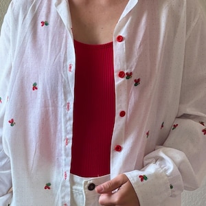 May include: A white button-down shirt with a red and green embroidered design. The shirt has a white polka dot pattern and is made of a lightweight fabric.