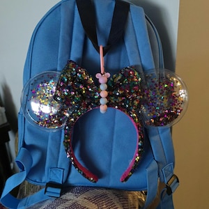 May include: A blue backpack with a sequined headband featuring a large bow and clear, round ears filled with colorful glitter. The headband has a pink interior and is adorned with a beaded charm.