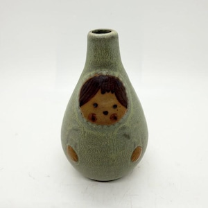 May include: A green ceramic vase with a child-like face design. The vase has a rounded body and a narrow neck. The face is brown with dark brown hair and small, round eyes. Two brown oval shapes are on the lower part of the vase.