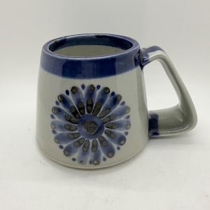 May include: A ceramic mug with a blue and grey floral design. The mug has a wide, rounded body with a blue rim and handle. The floral pattern is centered on the front of the mug.