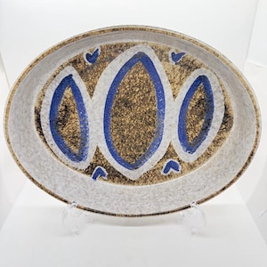 May include: Oval ceramic serving platter with a speckled brown centre, bordered by a white band and a speckled outer edge. The centre features three blue, stylized, oval shapes. Small blue heart-like accents are placed around the blue ovals.
