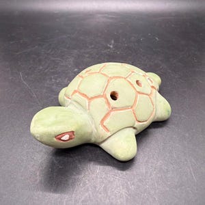 May include: A light green ceramic turtle ocarina featuring a smooth, matte finish. The shell is decorated with a raised, reddish-brown grid pattern and includes several circular finger holes. The head has a simple, stylized eye detail.