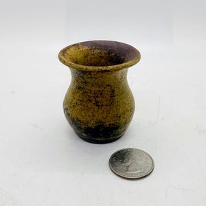 May include: A small, handcrafted ceramic vase with a rounded body and flared rim. The vase has a mottled, earthy colour palette of brown, yellow, and black. A coin is next to the vase for size comparison.