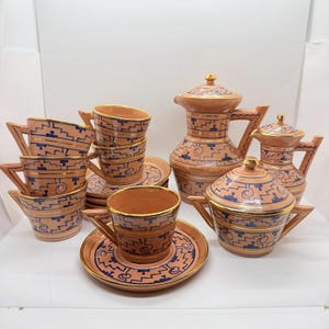 May include: A terracotta-coloured ceramic tea set with blue geometric patterns and gold trim. The set includes cups, saucers, a teapot, and a sugar bowl. The cups have angular handles, and the teapot has a lid with a gold finial.