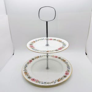 May include: Two-tiered serving stand with a silver metal handle and support. The white plates have a floral pattern with pink, blue, and yellow flowers, and a gold rim. Ideal for displaying desserts or appetizers.