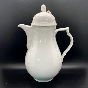 May include: White porcelain coffee pot with a lid and a floral design. The pot has a rounded body and a curved handle.