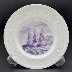May include: White ceramic plate with a purple illustration of sailing ships at sea. The plate has a detailed, embossed floral border. The image is set against a black background.
