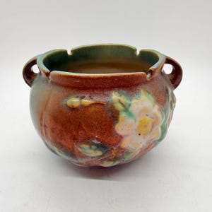 May include: A ceramic bowl with a brown exterior and a green interior rim. The bowl features a floral design with white, yellow, and green accents. It has two small handles on either side.