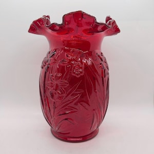 May include: A ruby red glass vase with a ruffled rim and embossed floral design. The vase has a bulbous shape and a detailed pattern of flowers and leaves. The glass is a deep, transparent red, and the vase is set against a white background.