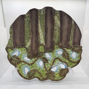 May include: A decorative ceramic plate with a forest scene. The top half features brown tree trunks with green accents, while the bottom half has green foliage with blue accents. The plate is supported by a clear stand.