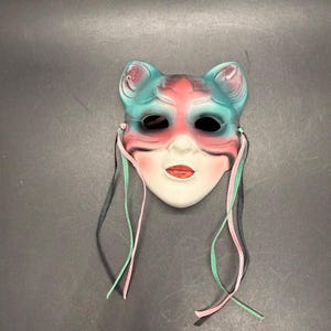 May include: A cat-shaped face mask featuring a gradient of teal and pink coloring across the forehead and cheeks. The mask has black eye cutouts, a white nose and chin area, red painted lips, and two thin ribbons in pink and teal attached to sides.