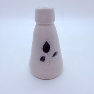 May include: A light pink ceramic bud vase with a dark purple floral design. The vase has a textured surface and a flared shape, with a cylindrical top. The floral design includes a leaf and two small buds.