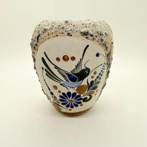 May include: A ceramic vase with a textured, light brown rim and a cream-coloured body. The vase features a hand-painted design of a bird, flowers, and foliage in blue, black, and brown. The vase has a unique, organic shape.