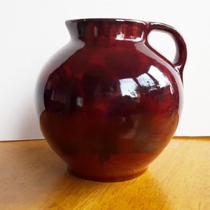 Può includere: Un'anfora in ceramica di colore rosso scuro con corpo arrotondato e un'unica maniglia. L'anfora ha una finitura lucida.