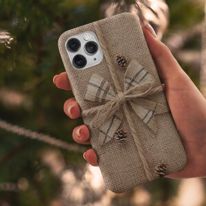May include: A beige phone case with a burlap texture, adorned with a plaid bow and twine. Small pinecones are attached to the case. The phone case is held in a hand.