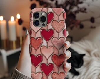 Puede incluir: Una funda de teléfono con un patrón de corazones en tonos rojos y rosas. La funda tiene un acabado brillante y un diseño de corazón repetido. El fondo está borroso, con velas y un gato negro visibles.