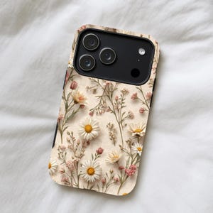 May include: A phone case featuring a floral design with white daisies, pink flowers, and green stems on a beige background. The case has a black border and a camera cutout.
