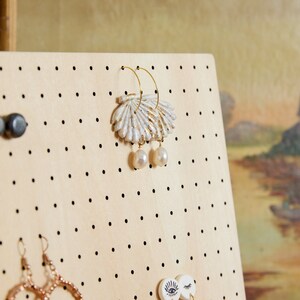May include: A wooden jewelry display board with black dots, featuring a pair of gold hoop earrings with a white and gold shell design and pearl accents, a pair of gold hoop earrings with a gold and white geometric design, and a pair of gold hoop earrings with a gold and white geometric design.
