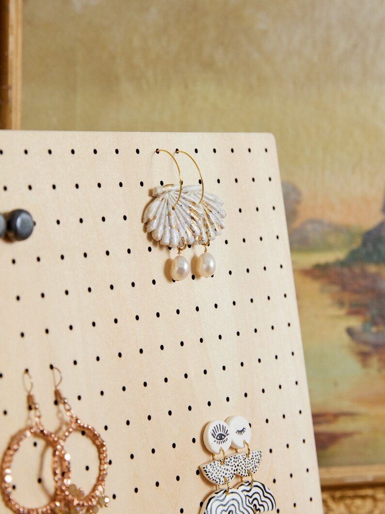 May include: A wooden jewelry display board with black dots, featuring a pair of gold hoop earrings with a white and gold shell design and pearl accents, a pair of gold hoop earrings with a gold and white geometric design, and a pair of gold hoop earrings with a gold and white geometric design.