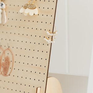 May include: A wooden earring display stand with a pegboard design and a curved base. The stand is made of light wood and has a small label that says "YOBI".
