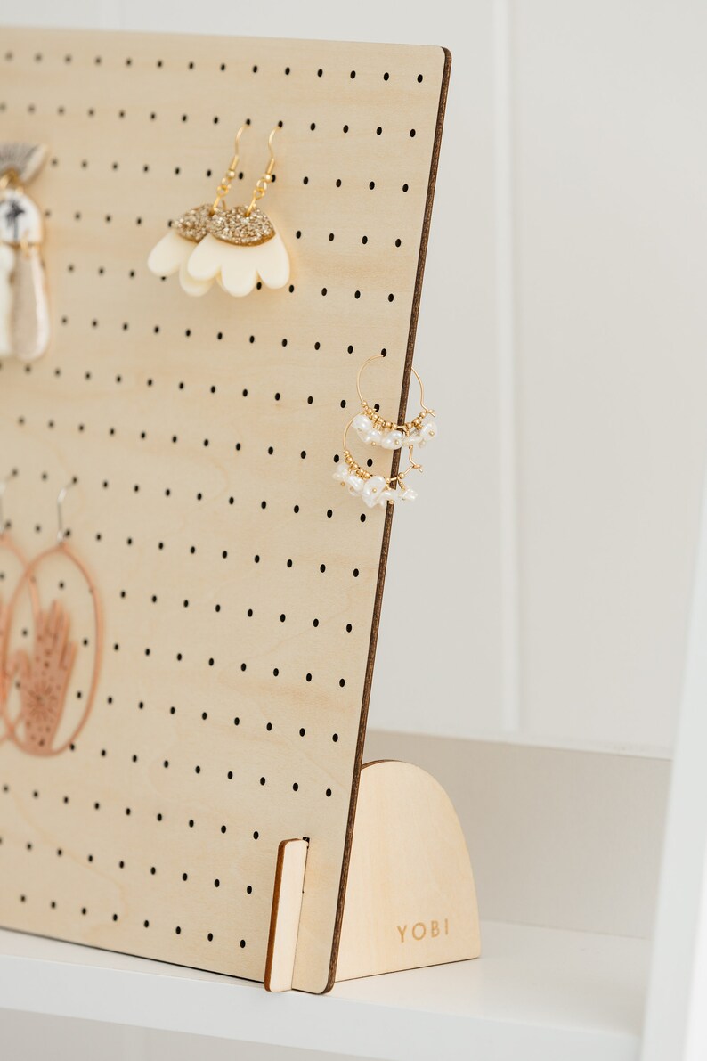 May include: A wooden earring display stand with a pegboard design and a curved base. The stand is made of light wood and has a small label that says "YOBI".