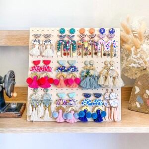 May include: A wooden display board with colorful, patterned earrings with tassels. The earrings are arranged in rows and are made of polymer clay. The earrings are all different shapes and sizes.