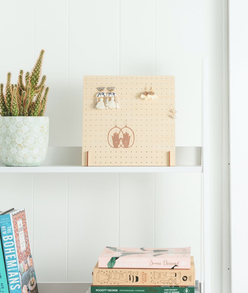 May include: A wooden earring display stand with a light brown finish. The stand has a grid of holes for hanging earrings and features a decorative design of two hands with a heart in the center. The stand is on a white shelf with books stacked below.