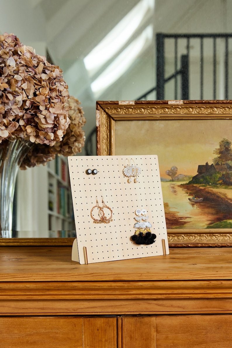 May include: A wooden earring display stand with a pegboard design. The stand has a variety of earrings hanging from it, including black tassel earrings, gold hoop earrings, and white shell earrings.