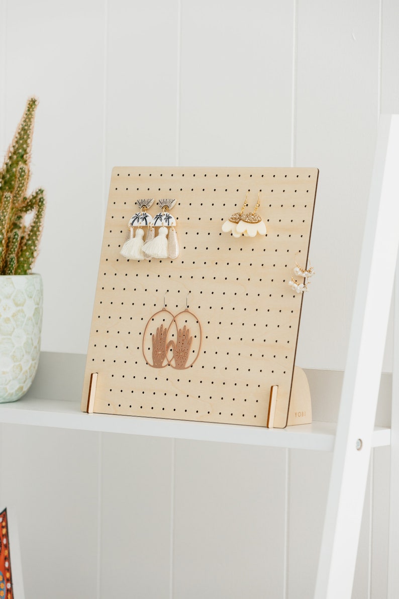 May include: A wooden earring display stand with a pegboard design. The stand has a light brown finish and is decorated with various earrings. The stand is labeled "YOBI" in the bottom right corner.