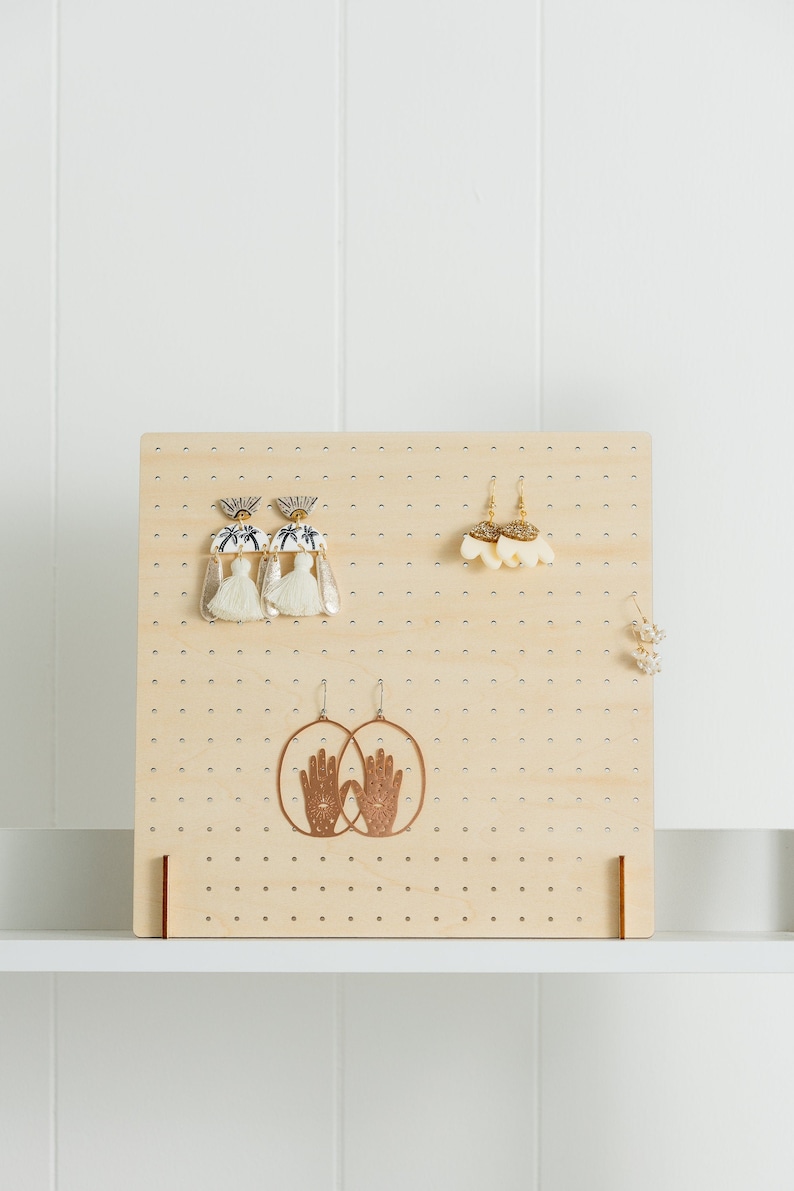 May include: A wooden earring display stand with a grid of holes. The stand has three pairs of earrings hanging from it, including a pair of white tassel earrings, a pair of gold shell earrings, and a pair of copper hand-shaped earrings.