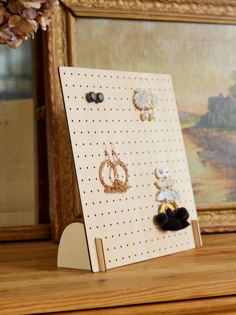 May include: A wooden earring holder with a pegboard design. The holder has a light brown finish and is standing on a wooden surface. There are several pairs of earrings hanging on the pegs.