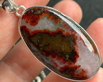 Op de afbeelding: Een zilveren hanger met een grote ovale agaat-steen met een uniek patroon in rood, wit en bruin. De steen is gezet in een zilveren bezel en hangt aan een zilveren ketting.