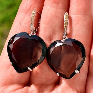 May include: A pair of heart-shaped earrings featuring large, faceted dark brown smoky quartz stones set in silver-toned metal. The top of each earring has a hinged clasp accented with small, clear, sparkling stones that catch the light.