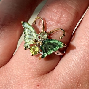 May include: A butterfly ring with green enamel wings and gold-colored accents. The ring features two round, light green gemstones and is set on a silver band. The butterfly's wings are detailed with dark green and light green enamel.