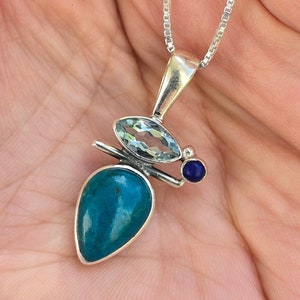 Op de afbeelding: Een zilveren ketting met een hanger met een traanvormige turquoise steen, een kleinere ronde lapis lazuli steen en een heldere aquamarijn steen.