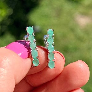 Puede incluir: Un par de pendientes de plata, cada uno con cinco esmeraldas ovaladas de color verde claro. Los pendientes se muestran sobre un fondo verde borroso, con una uña pintada de rosa en primer plano.