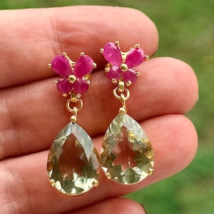 May include: A pair of gold-toned earrings featuring a cluster of pink gemstones at the top and a large, pear-shaped, light green gemstone dangling below. The earrings are displayed against a blurred green background.