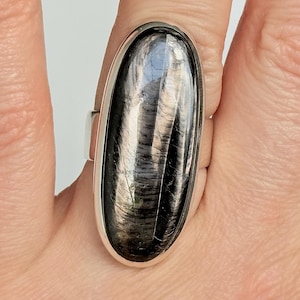 Peut inclure: Bague en argent avec une grosse pierre noire ovale. La pierre a un motif rayé réfléchissant. L'anneau est en argent poli et la pierre est sertie dans un sertissage à lunette.