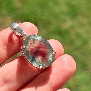 May include: Oval-shaped pendant with a large, translucent gemstone. The stone has a mix of green and red hues, set in a silver-colored metal frame. The pendant is suspended from a silver bail.
