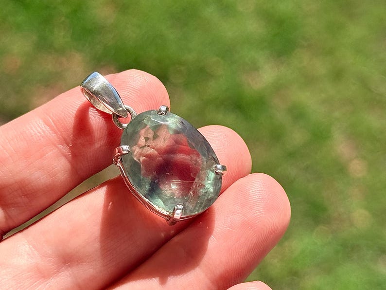 May include: Oval-shaped pendant with a large, translucent gemstone. The stone has a mix of green and red hues, set in a silver-colored metal frame. The pendant is suspended from a silver bail.