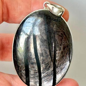 Pode incluir: Pingente oval com uma pedra cinza escuro polida com listras pretas verticais. A pedra é colocada em uma moldura de prata e tem uma fiança de prata. O pingente é segurado por uma mão.
