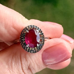 Op de afbeelding: Ovale robijnring met een halo van kleine, veelkleurige edelstenen. De band van de ring is zilverkleurig en de grote robijn heeft een dieprode kleur. De ring wordt door een hand omhoog gehouden.