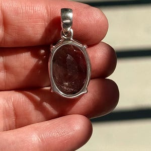 May include: A silver pendant with an oval, faceted, dark reddish-brown gemstone. The pendant has a simple, elegant design with a bail for a chain. The gemstone is set in a silver frame with small decorative elements.