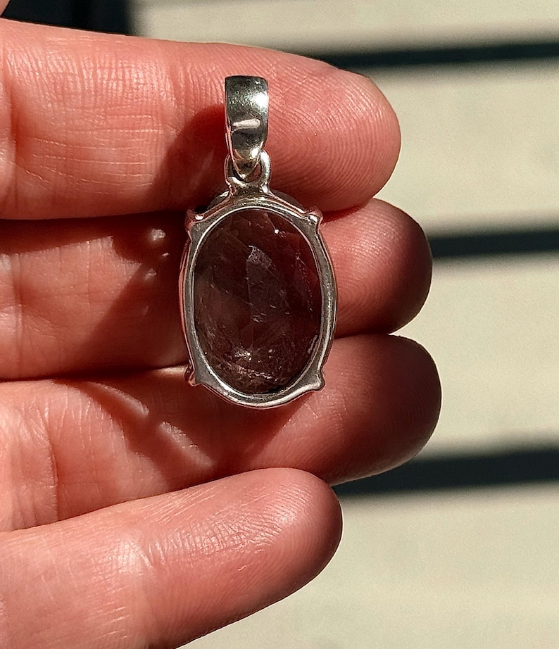 May include: A silver pendant with an oval, faceted, dark reddish-brown gemstone. The pendant has a simple, elegant design with a bail for a chain. The gemstone is set in a silver frame with small decorative elements.